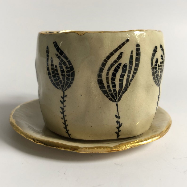 May include: A white ceramic teacup and saucer set with a black floral design and gold trim.