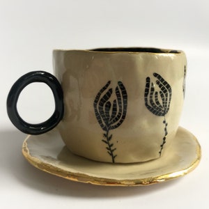 May include: A cream-colored ceramic teacup and saucer set with black floral designs. The saucer has a gold rim.