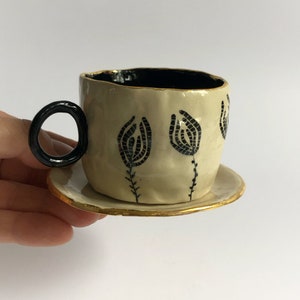 May include: A small, white ceramic teacup with a black handle and black floral design. The teacup has a gold rim and sits on a matching saucer.