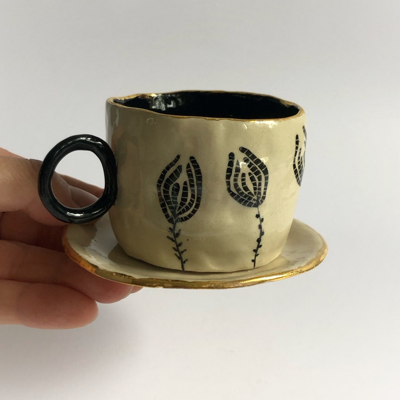May include: A small, white ceramic teacup with a black handle and black floral design. The teacup has a gold rim and sits on a matching saucer.