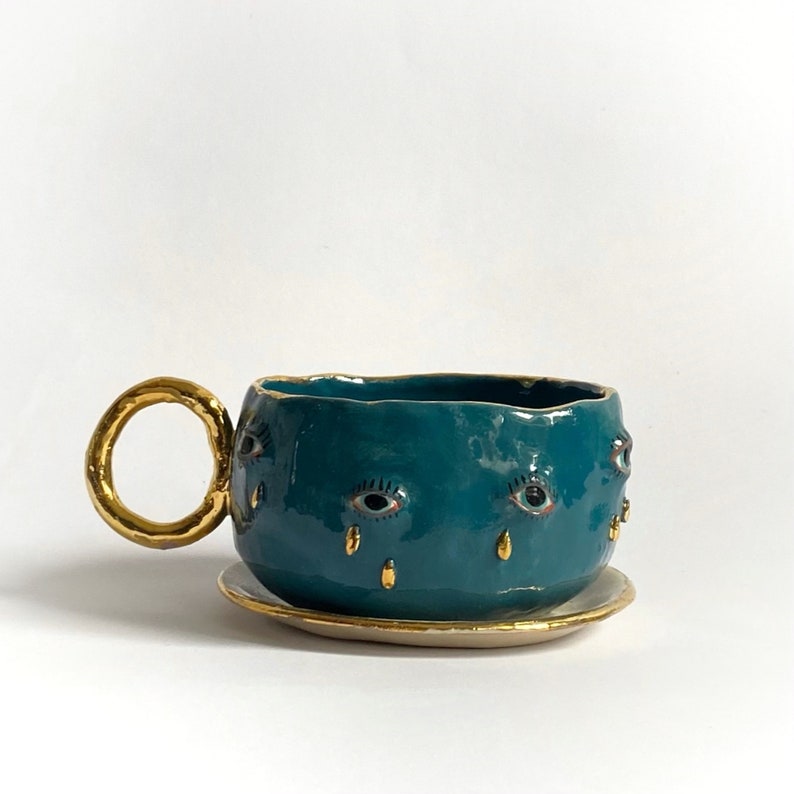 K&ouml;nnte beinhalten: Eine teeblau gef&auml;rbte Keramiktasse mit goldenem Henkel und Untertasse. Die Teetasse hat eine strukturierte Oberfl&auml;che und ist mit goldenen Augen verziert, die goldene Tr&auml;nen weinen.