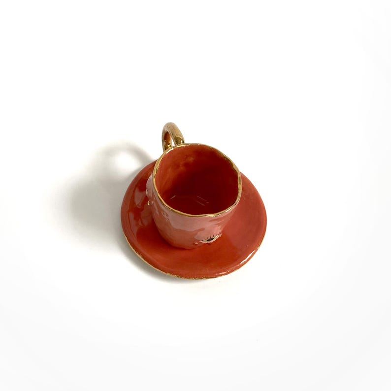 Puede incluir: Una taza de t&eacute; de cer&aacute;mica peque&ntilde;a de color coral con un borde dorado. La taza tiene una forma redondeada y una peque&ntilde;a asa.