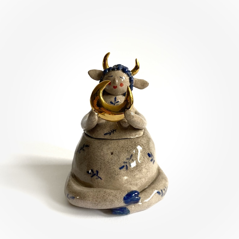 May include: A ceramic figurine of a seated cow holding a gold crescent moon. The cow has blue hair, gold horns, and blue floral accents on its body. The figurine is a light beige color with a matte finish.