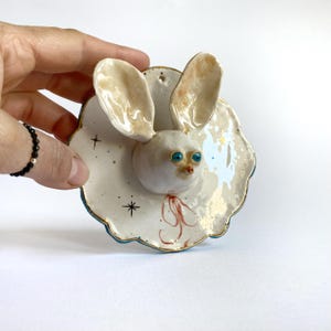 May include: A white ceramic wall hanging sculpture of a rabbit with large ears, blue eyes, and a red nose. The rabbit is attached to a white ceramic plate with a blue rim and gold accents. The plate has a star pattern and a light blue and white splatter paint design.