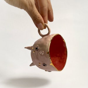 May include: A pink ceramic cup with a gold rim and a red interior. The cup has a handle and is decorated with gold and black eyes.