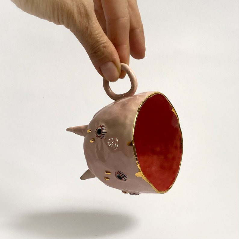 May include: A pink ceramic cup with a gold rim and a red interior. The cup has a handle and is decorated with gold and black eyes.