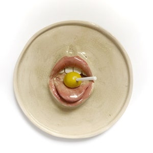 May include: A ceramic plate featuring a 3D sculpted mouth with a pink tongue and lips, holding a yellow lollipop on a white stick.