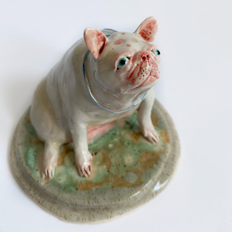 May include: A ceramic French Bulldog figurine with a light pink and white coat, featuring blue details. The dog sits on a round, green and brown base. The dog's face has pink spots and blue eyes.