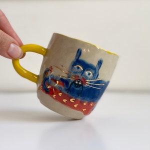 Puede incluir: Una taza de cer&aacute;mica con asa amarilla y un gato azul con bigotes dorados comiendo una rana negra. El gato est&aacute; sobre un fondo estampado rojo y dorado.
