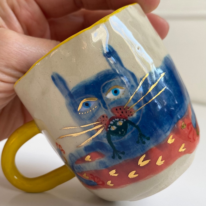 Puede incluir: Una taza de cer&aacute;mica amarilla con una cara de gato azul y detalles dorados. El gato tiene una boca roja y una rana verde en su boca. La taza tiene un patr&oacute;n rojo y dorado en la parte inferior.