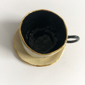 May include: A white ceramic teacup and saucer with a black interior and gold trim. The cup has a unique, slightly irregular shape.