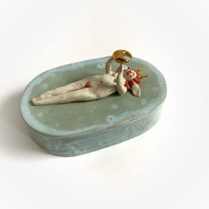 May include: A ceramic box with a light blue glaze features a sculpted figure of a nude woman with red hair and horns, holding a gold crescent moon. The box has a white interior and a textured surface.