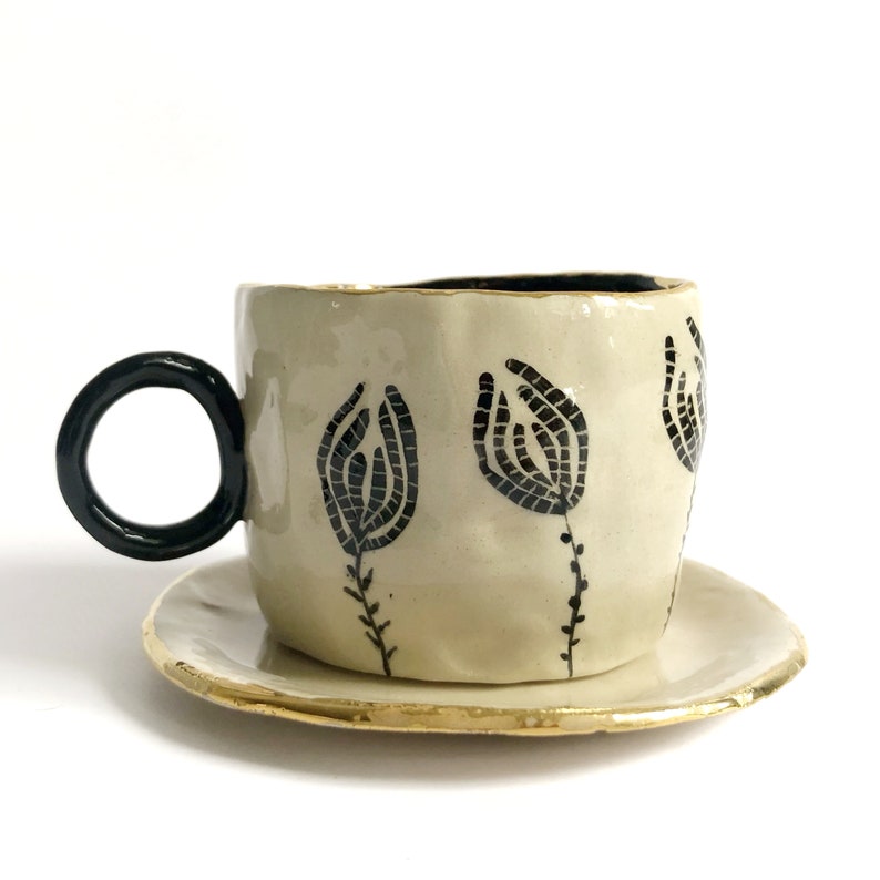 May include: A white ceramic teacup and saucer with a black floral design. The saucer has a gold rim.