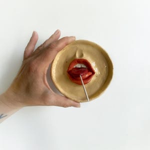 Könnte beinhalten: Eine Keramikschale mit einem Gesichtsdesign, das rote Lippen und eine Zunge mit einem Lutscher zeigt. Die Schale wird von einer Hand vor einem weißen Hintergrund gehalten. Die Schale ist beige.