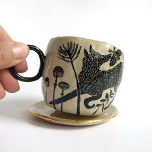 Peut inclure: Une tasse à thé et une soucoupe en céramique avec une illustration en noir et blanc d'un renard, de champignons et d'un papillon de nuit. La soucoupe a un bord doré.