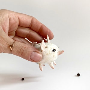 Puede incluir: Una figurita de cerdo de cer&aacute;mica blanca con ojos negros y una nariz rosa. El cerdo est&aacute; de pie sobre sus cuatro patas y tiene una textura punteada.
