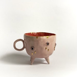 May include: A pink ceramic mug with three legs and gold accents. The mug has a red interior and features three eyes with gold eyelashes and tears.
