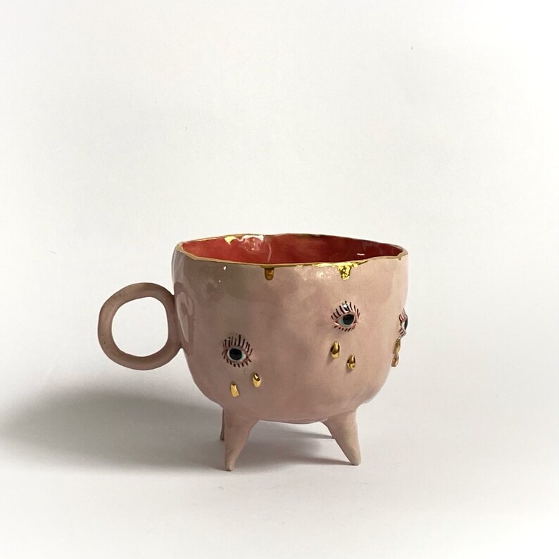 May include: A pink ceramic mug with three legs and gold accents. The mug has a red interior and features three eyes with gold eyelashes and tears.