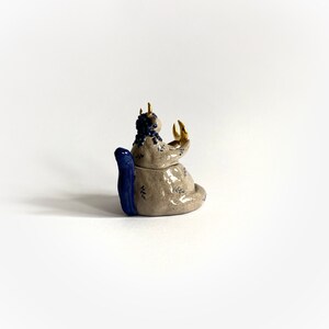 May include: A ceramic sculpture of a seated creature with a blue tail and blue hair. The creature has gold horns and is holding its hands up. The sculpture is beige with blue and gold accents and leaf patterns.