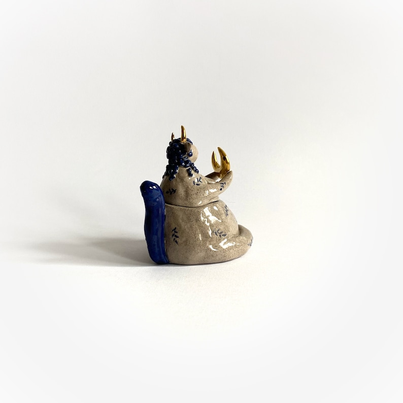 May include: A ceramic sculpture of a seated creature with a blue tail and blue hair. The creature has gold horns and is holding its hands up. The sculpture is beige with blue and gold accents and leaf patterns.
