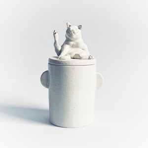 Peut inclure: Pot en céramique blanche avec un couvercle en forme de chat assis sur le dessus. Le chat a sa patte levée et regarde sur le côté.