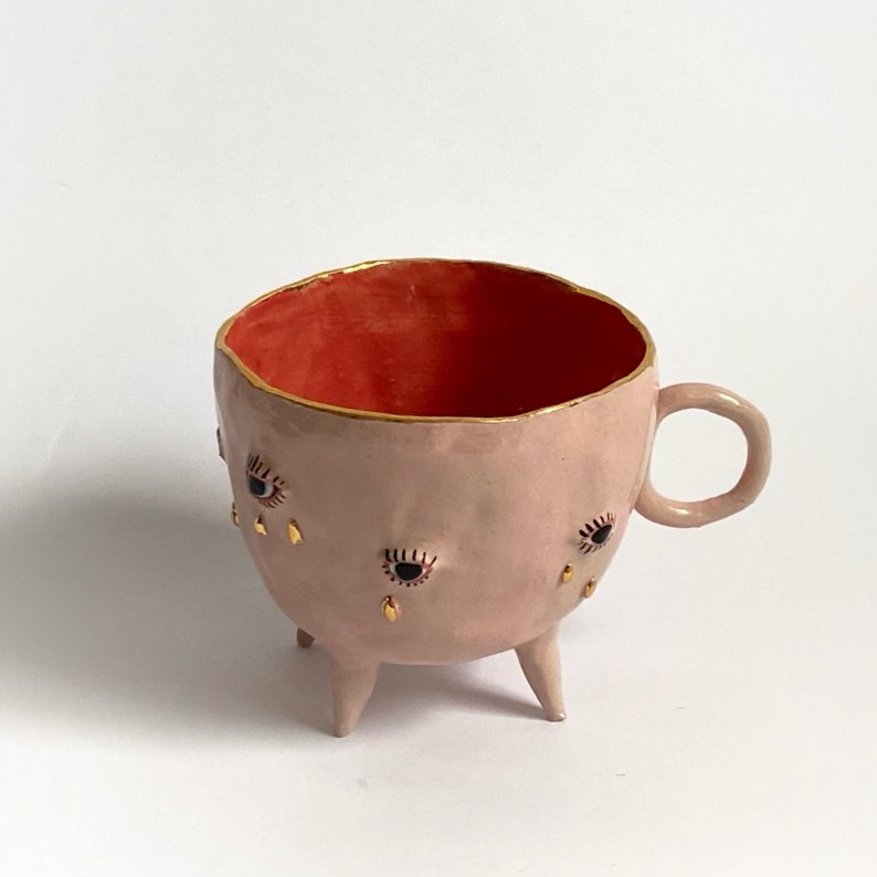May include: A pink ceramic mug with three legs and a handle. The mug is decorated with gold accents and black and red eyes that have gold teardrops.