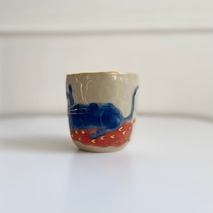 Puede incluir: Una taza de cer&aacute;mica con un dise&ntilde;o de gato azul. El gato est&aacute; tumbado sobre un coj&iacute;n con estampado rojo y dorado. La taza tiene un borde amarillo.