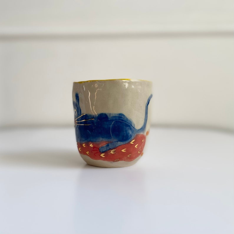 Puede incluir: Una taza de cer&aacute;mica con un dise&ntilde;o de gato azul. El gato est&aacute; tumbado sobre un coj&iacute;n con estampado rojo y dorado. La taza tiene un borde amarillo.