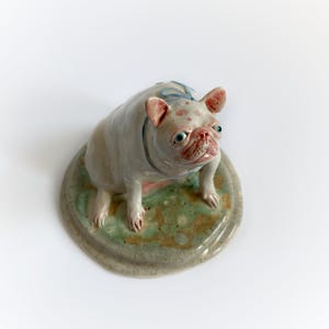 May include: A ceramic pug dog figurine with a light pink face and ears, and a light blue collar. The dog stands on a round, mottled green and brown base. The dog's body is white with pink spots.
