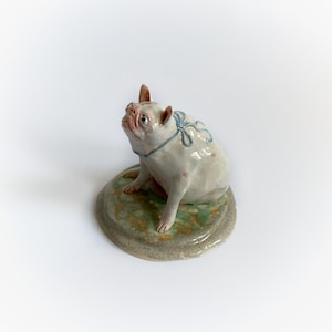 May include: A ceramic bulldog figurine, white with blue ribbon detail, sits on a round, mottled green and grey base. The dog has pink spots and an upturned snout. A decorative item.