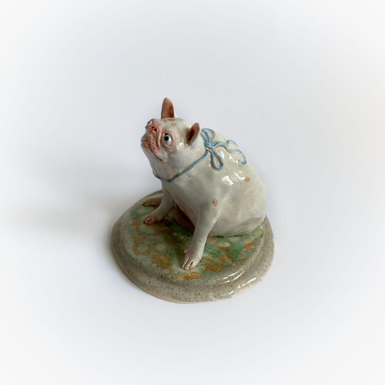 May include: A ceramic bulldog figurine, white with blue ribbon detail, sits on a round, mottled green and grey base. The dog has pink spots and an upturned snout. A decorative item.