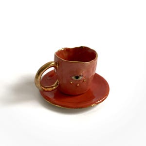 Puede incluir: Un juego de taza y platillo de t&eacute; de cer&aacute;mica roja peque&ntilde;a con borde dorado. La taza de t&eacute; tiene un asa dorada y un ojo pintado con detalles dorados y una sola l&aacute;grima.