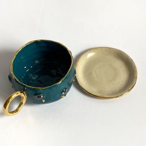 K&ouml;nnte beinhalten: Teetasse aus blaugr&uuml;ner Keramik mit goldener Verzierung und schwarzen Augendetails. Die Teetasse hat eine passende Untertasse.