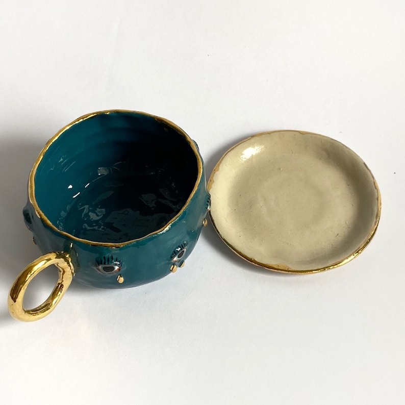 K&ouml;nnte beinhalten: Teetasse aus blaugr&uuml;ner Keramik mit goldener Verzierung und schwarzen Augendetails. Die Teetasse hat eine passende Untertasse.