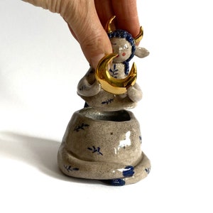 May include: A ceramic figurine of a seated figure with blue hair and gold horns, holding a gold crescent moon. The figure is in a small, textured pot with blue leaf designs. The overall color palette is neutral with blue and gold accents.