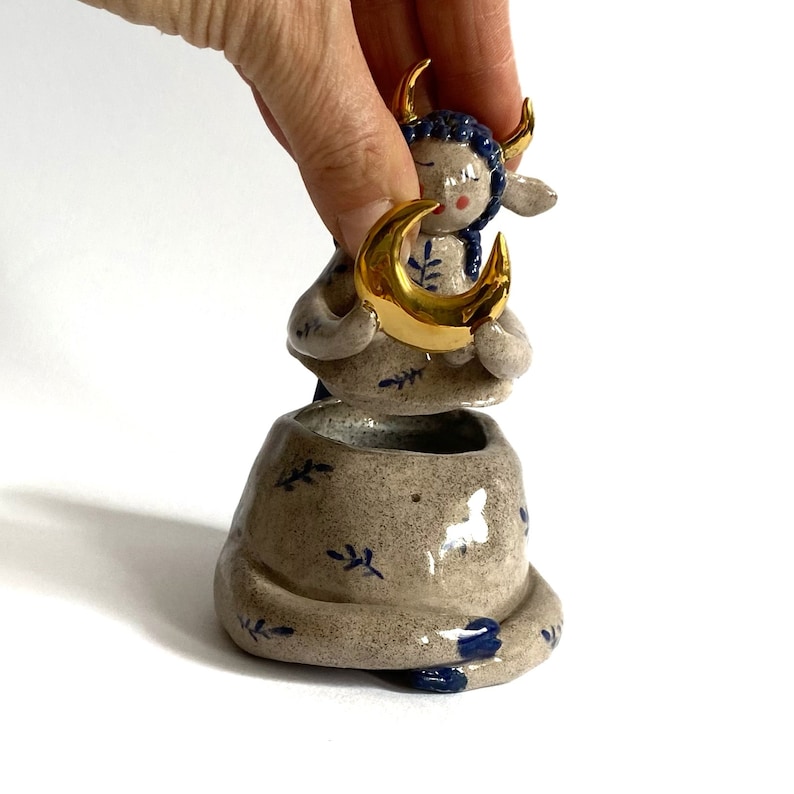 May include: A ceramic figurine of a seated figure with blue hair and gold horns, holding a gold crescent moon. The figure is in a small, textured pot with blue leaf designs. The overall color palette is neutral with blue and gold accents.