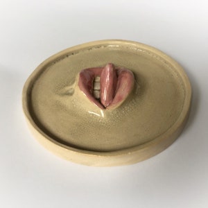 Pode incluir: Um prato de cer&acirc;mica com uma imagem esculpida em relevo de uma boca com l&aacute;bios rosados e uma l&iacute;ngua que se projeta.