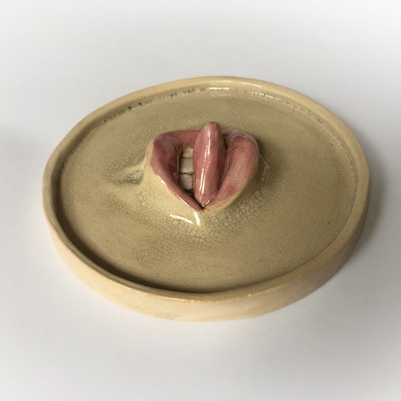 Pode incluir: Um prato de cer&acirc;mica com uma imagem esculpida em relevo de uma boca com l&aacute;bios rosados e uma l&iacute;ngua que se projeta.