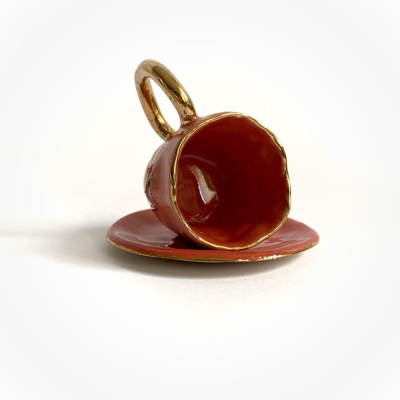 Puede incluir: Una peque&ntilde;a taza de t&eacute; de cer&aacute;mica roja con un borde y un asa dorados, sentada boca abajo sobre un platillo a juego.