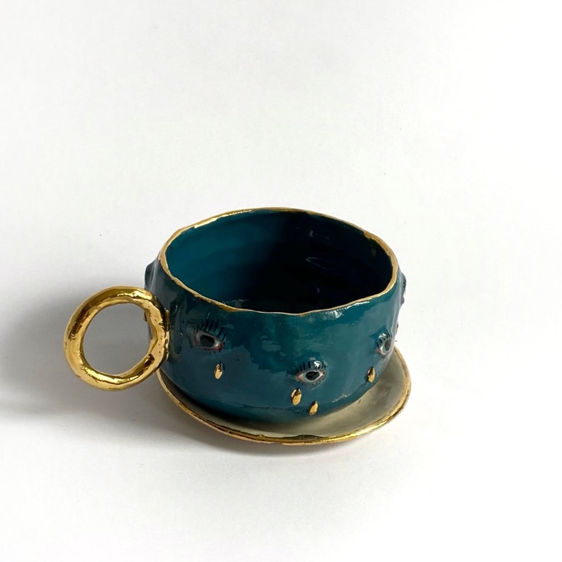 K&ouml;nnte beinhalten: Eine teeblau gef&auml;rbte Keramiktasse mit goldfarbenem Rand und Untertasse. Die Teetasse hat drei Augen mit goldenen Wimpern und goldenen Tr&auml;nentropfen-Details.