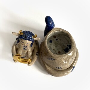 May include: Handmade ceramic art piece featuring a seated figure with blue hair and gold horns, holding a gold crescent. Beside it is a tan and blue ceramic vessel with a blue accent.