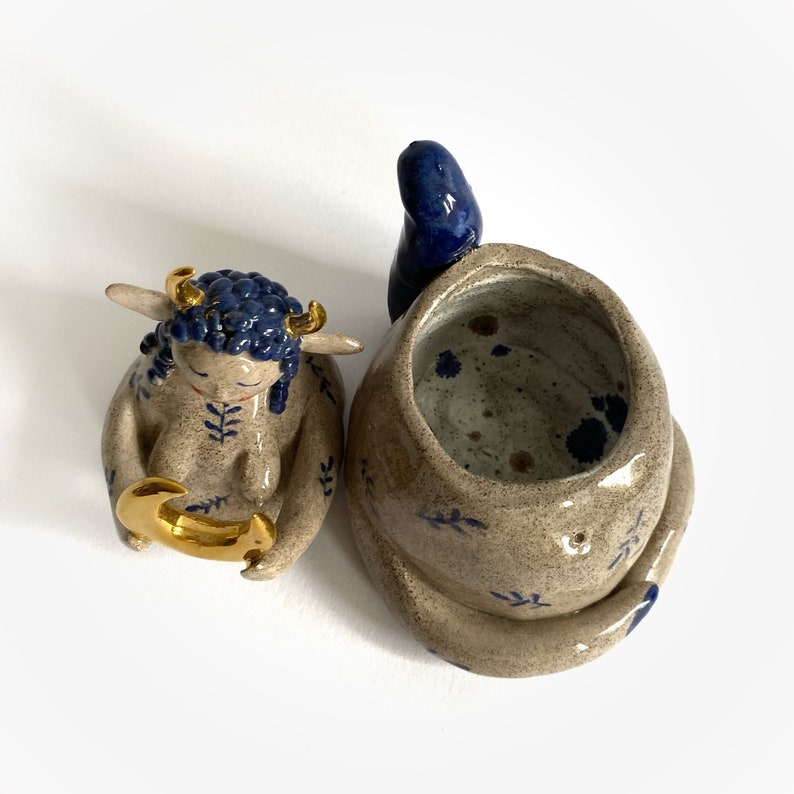 May include: Handmade ceramic art piece featuring a seated figure with blue hair and gold horns, holding a gold crescent. Beside it is a tan and blue ceramic vessel with a blue accent.