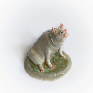 May include: A ceramic bulldog sculpture in light grey, with pink ears and a blue collar. It sits on a round base with green and tan mottled patterns. The dog is looking upwards. The sculpture is a decorative item.