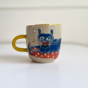 Puede incluir: Una taza de cer&aacute;mica con asa amarilla y un gato azul con detalles rojos y dorados pintados en el lateral.