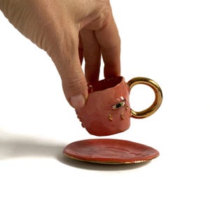 Puede incluir: Una taza de t&eacute; de cer&aacute;mica rosa con asa dorada y un dise&ntilde;o de ojo &uacute;nico. La taza est&aacute; sobre un platillo rosa a juego con un borde dorado.