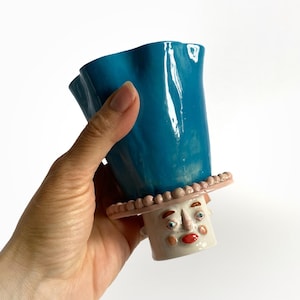 Op de afbeelding: Een grillige keramische mok met een turquoise blauwe, gerimpelde bovenkant en een gezichtvormige basis. De basis heeft een wit gezicht met rode lippen, blauwe ogen en een roze hoed met kleine, ronde versieringen. De mok wordt door een hand vastgehouden.