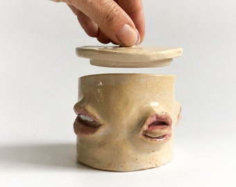 May include: A tan ceramic jar with a lid, featuring two sets of lips as a design element. The jar is held open by a hand, revealing the interior. The piece has a unique, artistic aesthetic.