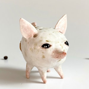 Puede incluir: Figurita de cer&aacute;mica blanca de un cerdo con ojos negros y nariz rosa. El cerdo tiene un esmalte blanco y marr&oacute;n moteado y se para sobre cuatro patas cortas.