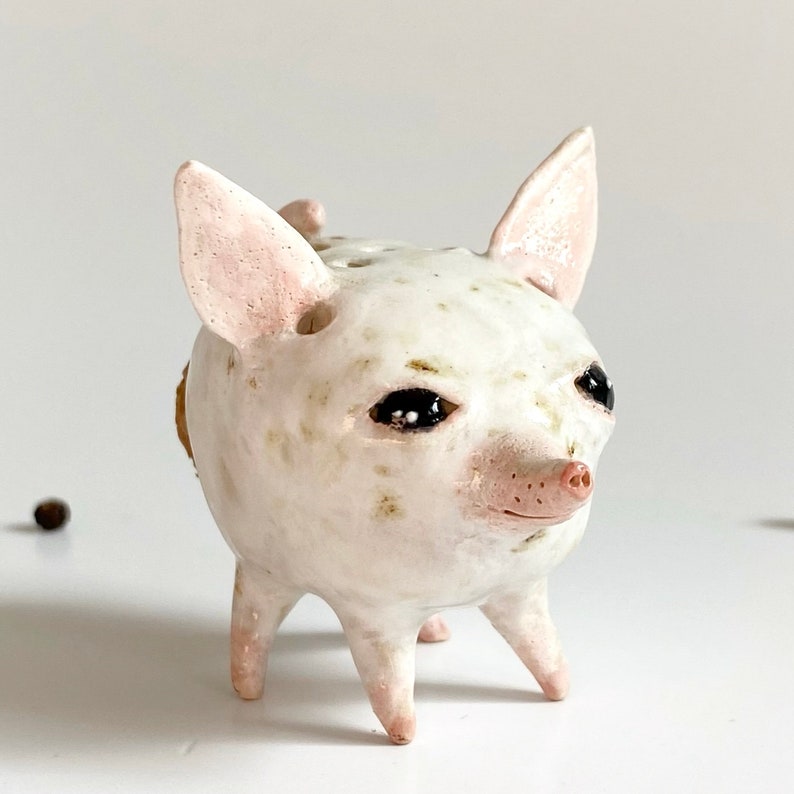 Puede incluir: Figurita de cer&aacute;mica blanca de un cerdo con ojos negros y nariz rosa. El cerdo tiene un esmalte blanco y marr&oacute;n moteado y se para sobre cuatro patas cortas.