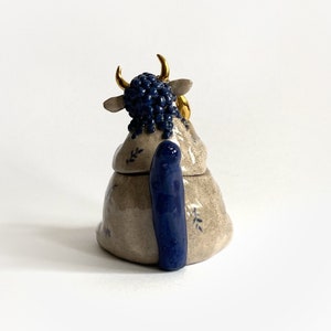 May include: A ceramic cow-shaped container with a lid. The cow has a blue, textured head and tail, with gold-colored horns and ear accents. The body is beige with blue floral accents.